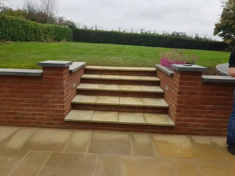 A new back Garden wall of red bricks leading on newly made steps. New yellow patio flooring on ground and on the steps. Top of brick with  greyish coloured tile.