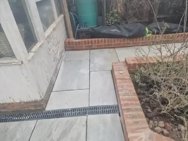 image of back garden , grey indian sandstone patio with a tiny wall of height three bricks around the edges of the patio. Drainage added to stop flooding.